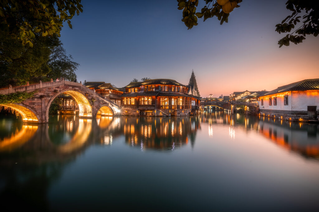 Eine beleuchtete Steinbrücke mit Rundbögen und traditionellen Häusern spiegelt sich im Wasser eines Kanals bei Sonnenuntergang, der Himmel zeigt Farbverläufe von Blau bis Orange.